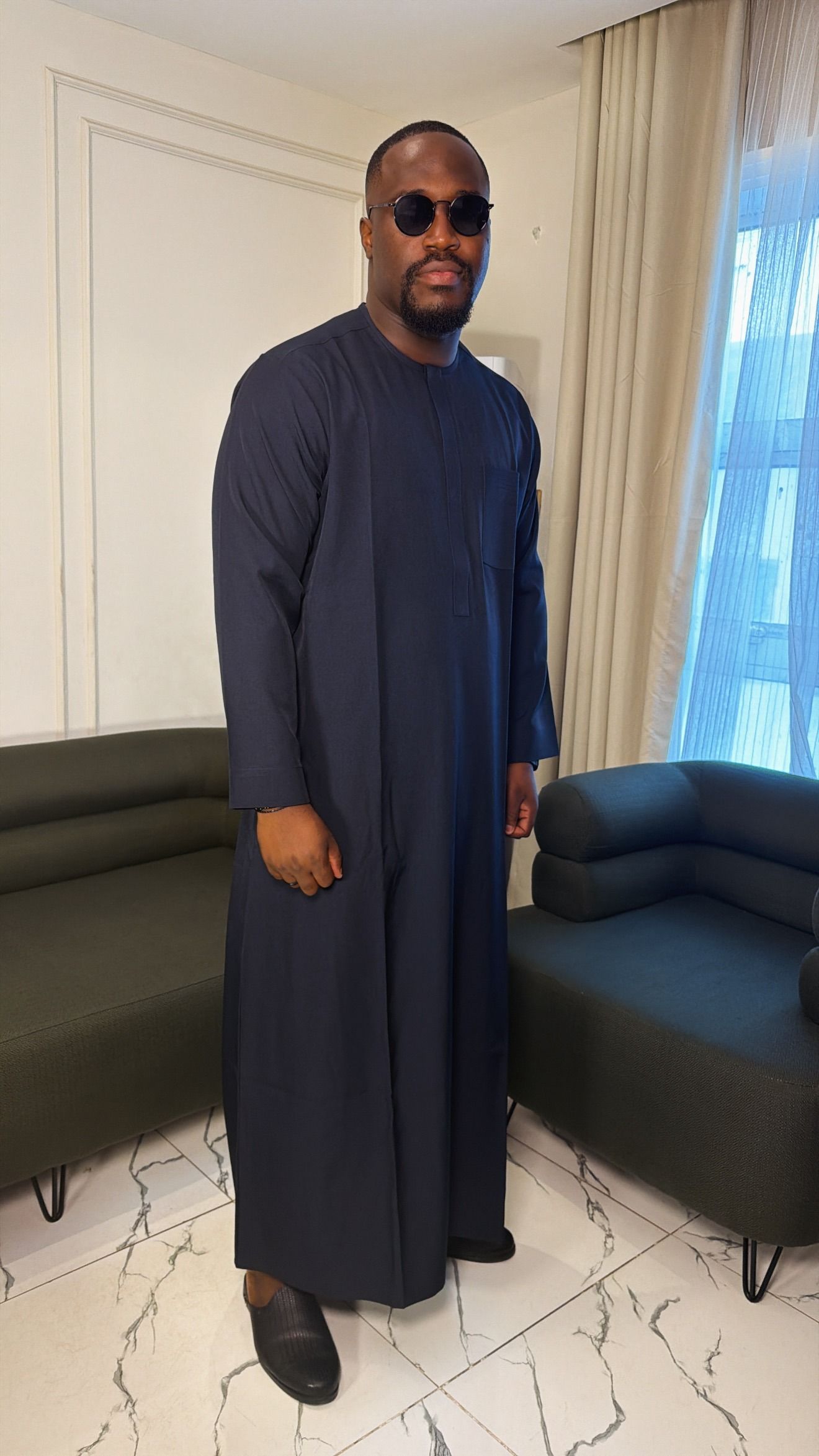 Man in a full-length navy kaftan standing by a window