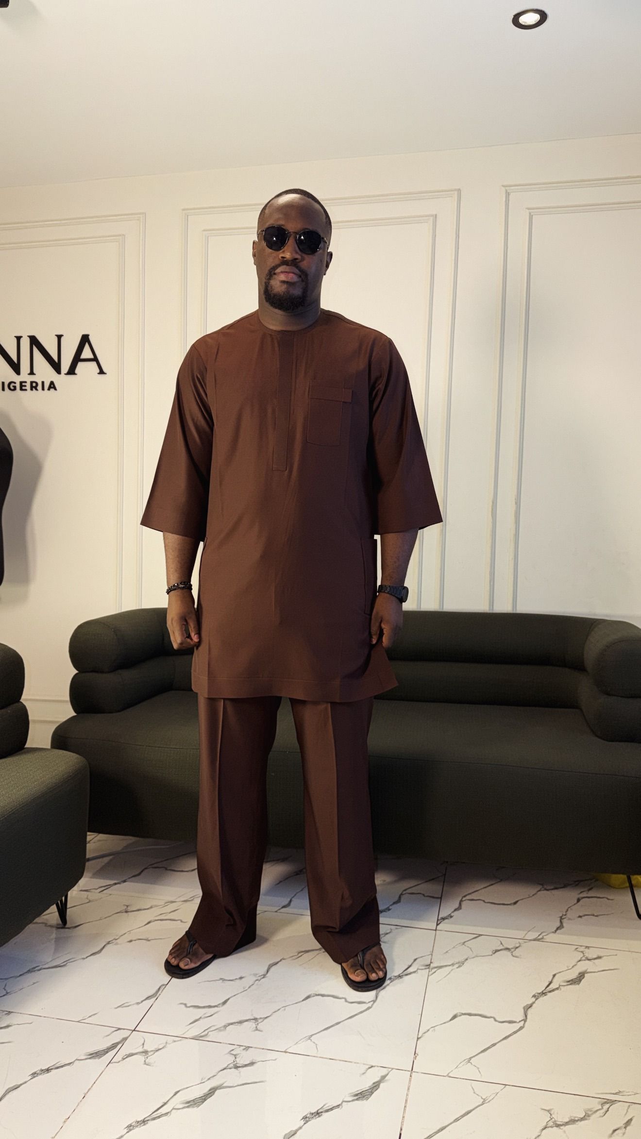 Man in a chocolate brown tunic, three-quarter view beside the JINNA sign